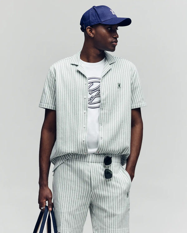 A man in a Psycho Bunny MENS ROBERT TEXTURE STRIPE SHIRT (MT0300005), matching shorts, blue cap, graphic tee, striped tote, and sunglasses clipped to his shorts stands against a plain background.