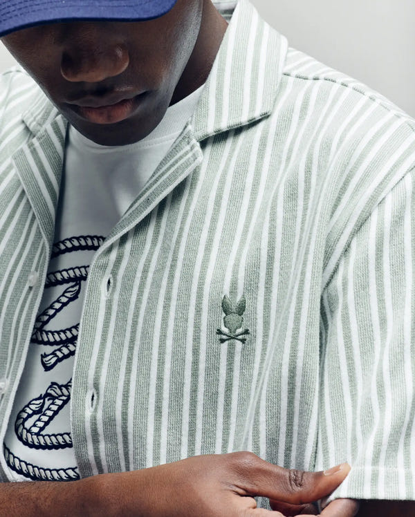 A person wears the Psycho Bunny MENS ROBERT TEXTURE STRIPE SHIRT (MT0300005), 100% cotton with embroidered logo and green-white stripes, layered over a white T-shirt with a rope design; focus is on clothing details, not the face.