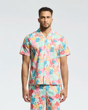 A person in Psycho Bunny's MENS MEYLON STRETCH POPLIN SHIRT (MT0300034), a short-sleeve, multi-colored floral with an embroidered logo, matching pants, camp collar, and vibrant pink, orange, blue, and green pattern stands against a plain background.