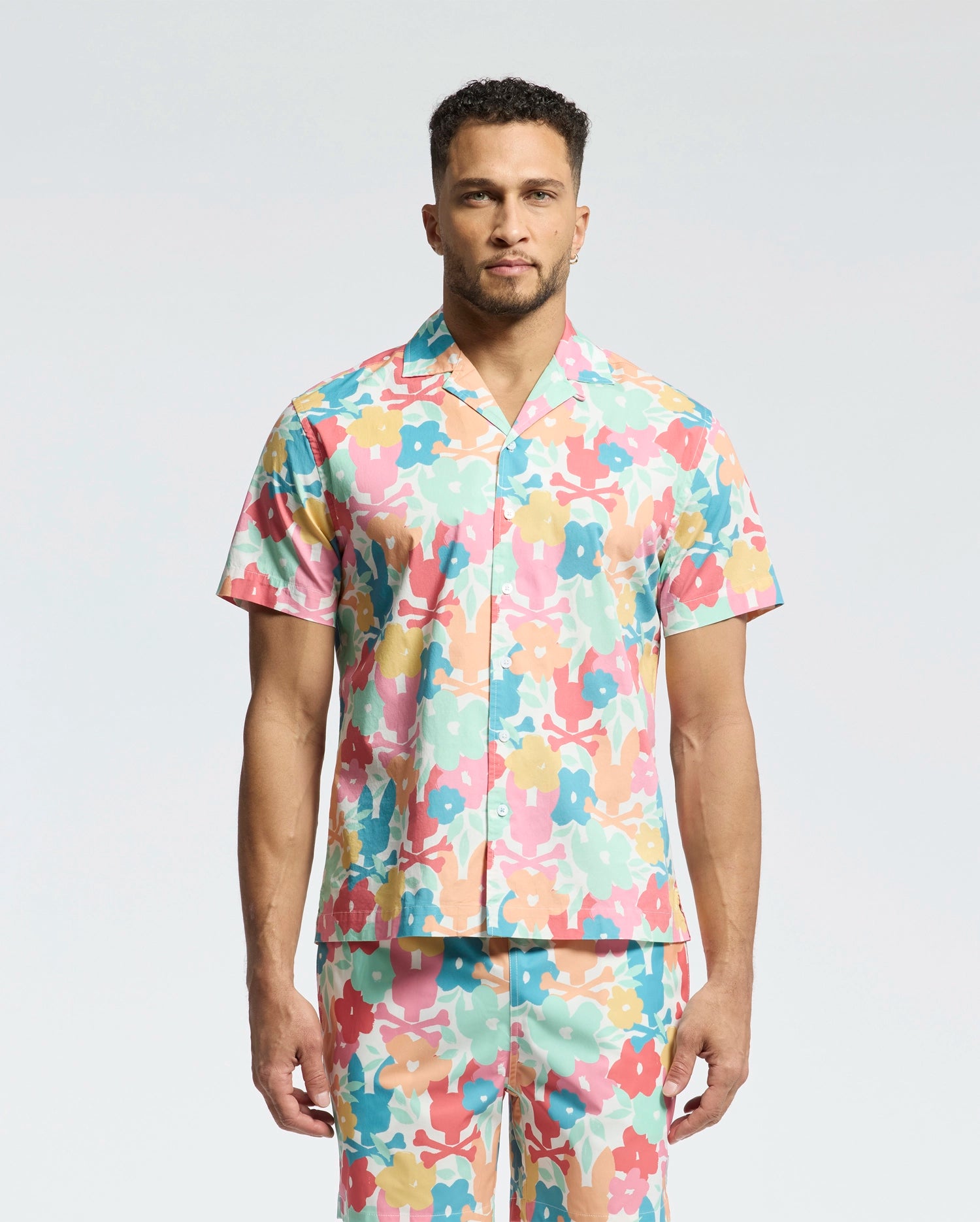 A person in Psycho Bunny's MENS MEYLON STRETCH POPLIN SHIRT (MT0300034), a short-sleeve, multi-colored floral with an embroidered logo, matching pants, camp collar, and vibrant pink, orange, blue, and green pattern stands against a plain background.