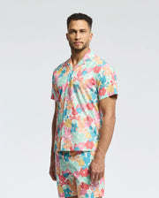 A man stands confidently against a plain background wearing the Psycho Bunny MENS MEYLON STRETCH POPLIN SHIRT (MT0300034) and matching pants. The ensemble features a vibrant floral pattern in pink, green, orange, and blue with a camp collar that complements his short curly hair and beard.