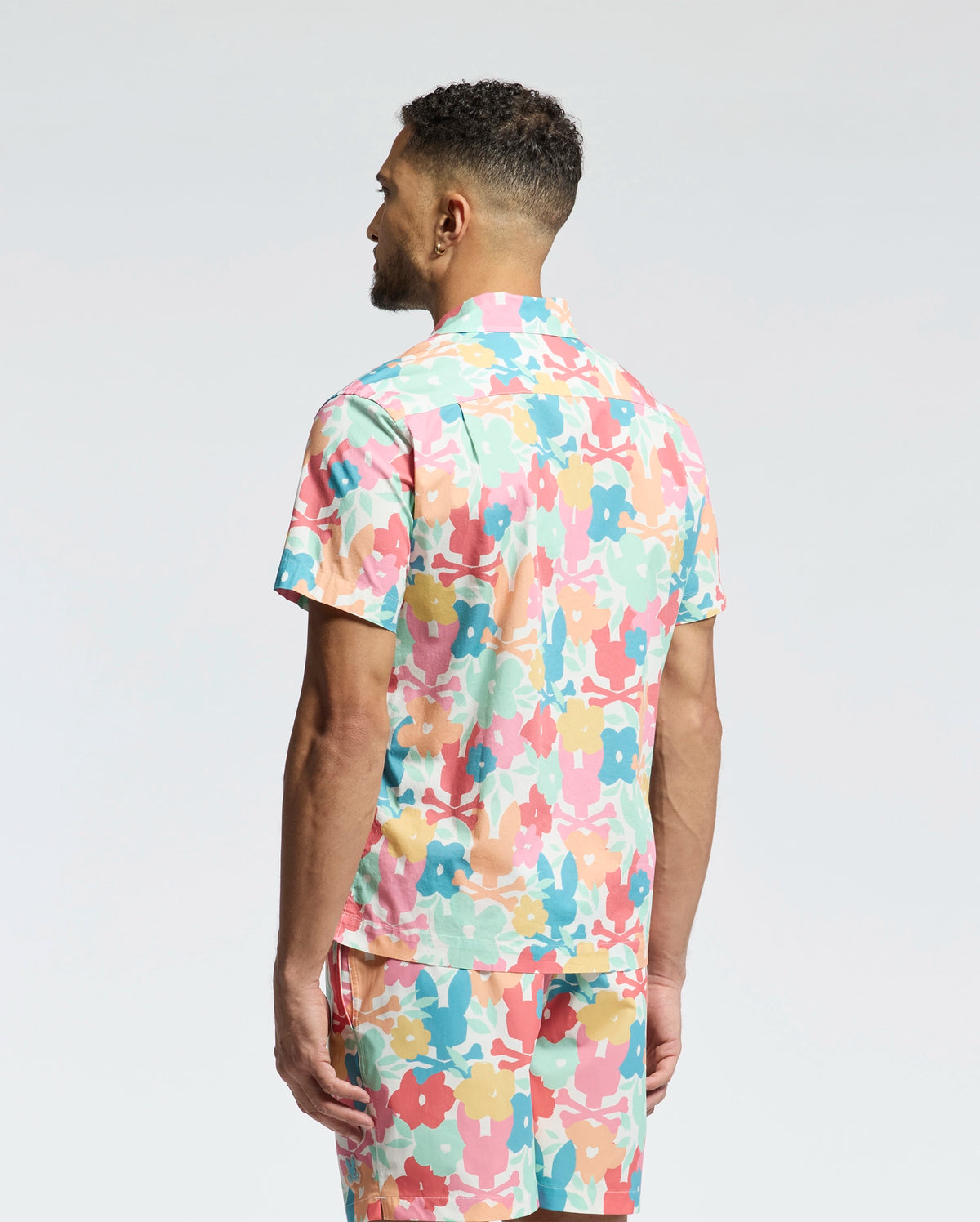 A man with short curly hair stands with his back to the camera, wearing Psycho Bunny's MENS MEYLON STRETCH POPLIN SHIRT (MT0300034) featuring a colorful floral camp collar and matching shorts against a plain white background.