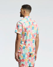 A man with short curly hair stands with his back to the camera, wearing Psycho Bunny's MENS MEYLON STRETCH POPLIN SHIRT (MT0300034) featuring a colorful floral camp collar and matching shorts against a plain white background.