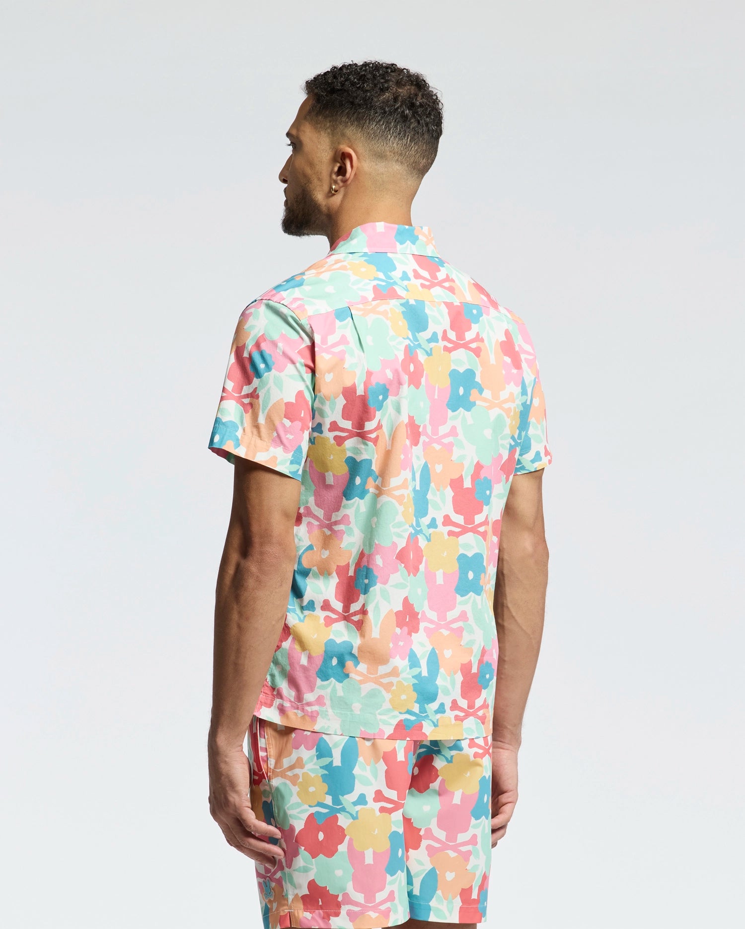 A man with short curly hair stands with his back to the camera, wearing Psycho Bunny's MENS MEYLON STRETCH POPLIN SHIRT (MT0300034) featuring a colorful floral camp collar and matching shorts against a plain white background.