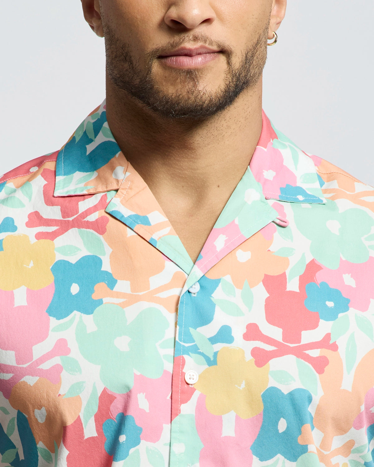 A bearded individual with a hoop earring dons the Psycho Bunny MENS MEYLON STRETCH POPLIN SHIRT (MT0300034), featuring colorful floral shades of pink, blue, yellow, and peach. The relaxed vibe comes from its camp collar and button-down style, subtly accented by an embroidered logo against a plain light background.