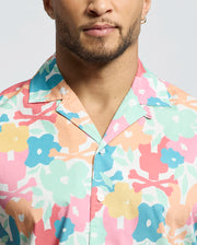 A bearded individual with a hoop earring dons the Psycho Bunny MENS MEYLON STRETCH POPLIN SHIRT (MT0300034), featuring colorful floral shades of pink, blue, yellow, and peach. The relaxed vibe comes from its camp collar and button-down style, subtly accented by an embroidered logo against a plain light background.