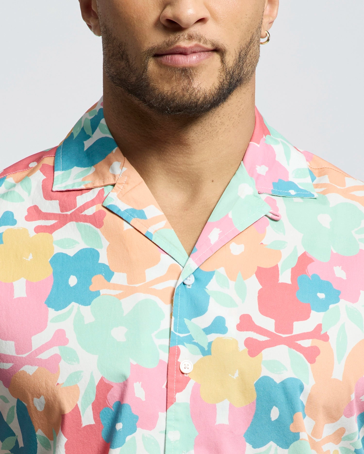 A bearded individual with a hoop earring dons the Psycho Bunny MENS MEYLON STRETCH POPLIN SHIRT (MT0300034), featuring colorful floral shades of pink, blue, yellow, and peach. The relaxed vibe comes from its camp collar and button-down style, subtly accented by an embroidered logo against a plain light background.