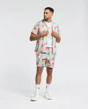 A man wears a Psycho Bunny MENS MEYLON STRETCH POPLIN SHIRT (MT0300034) with matching shorts, adorned in abstract patterns and a camp collar. With a white t-shirt beneath, white socks, and sneakers, he stands against a plain background, hands on his open shirt, looking to the side.