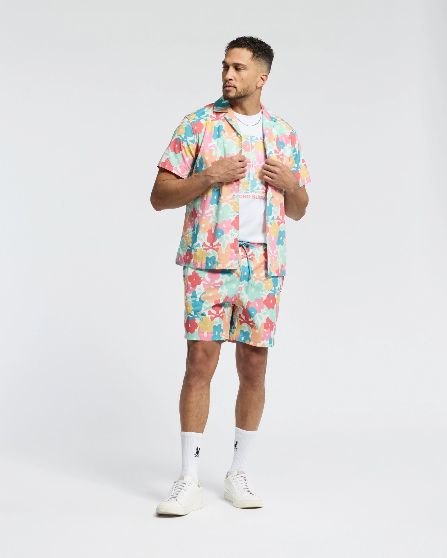 A man wears a Psycho Bunny MENS MEYLON STRETCH POPLIN SHIRT (MT0300034) with matching shorts, adorned in abstract patterns and a camp collar. With a white t-shirt beneath, white socks, and sneakers, he stands against a plain background, hands on his open shirt, looking to the side.