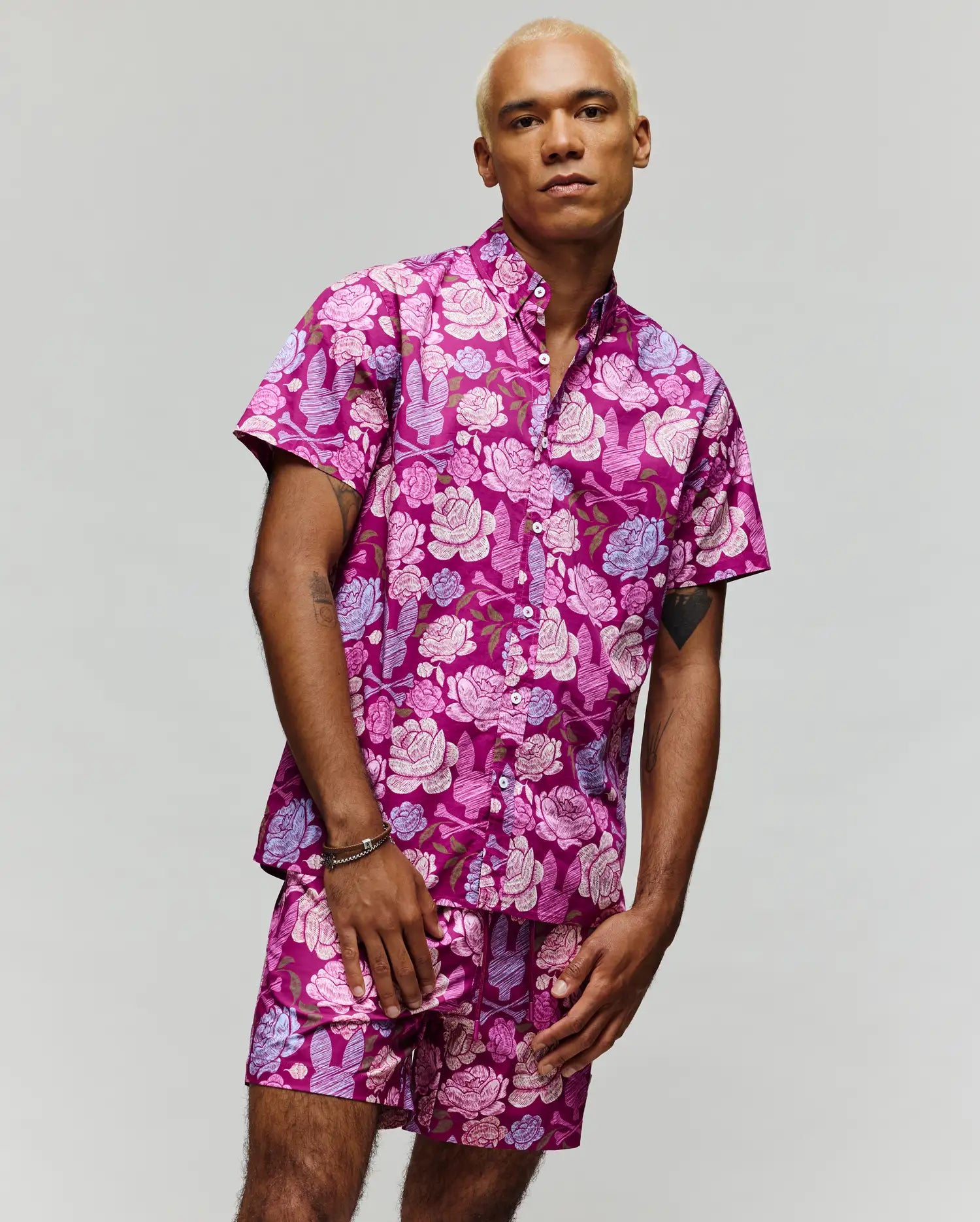 A man with short blond hair wears the Psycho Bunny MENS ROWEN SHIRT - MT0300038, a cotton blend with a vibrant pink and purple rose floral pattern, standing against a plain light background.