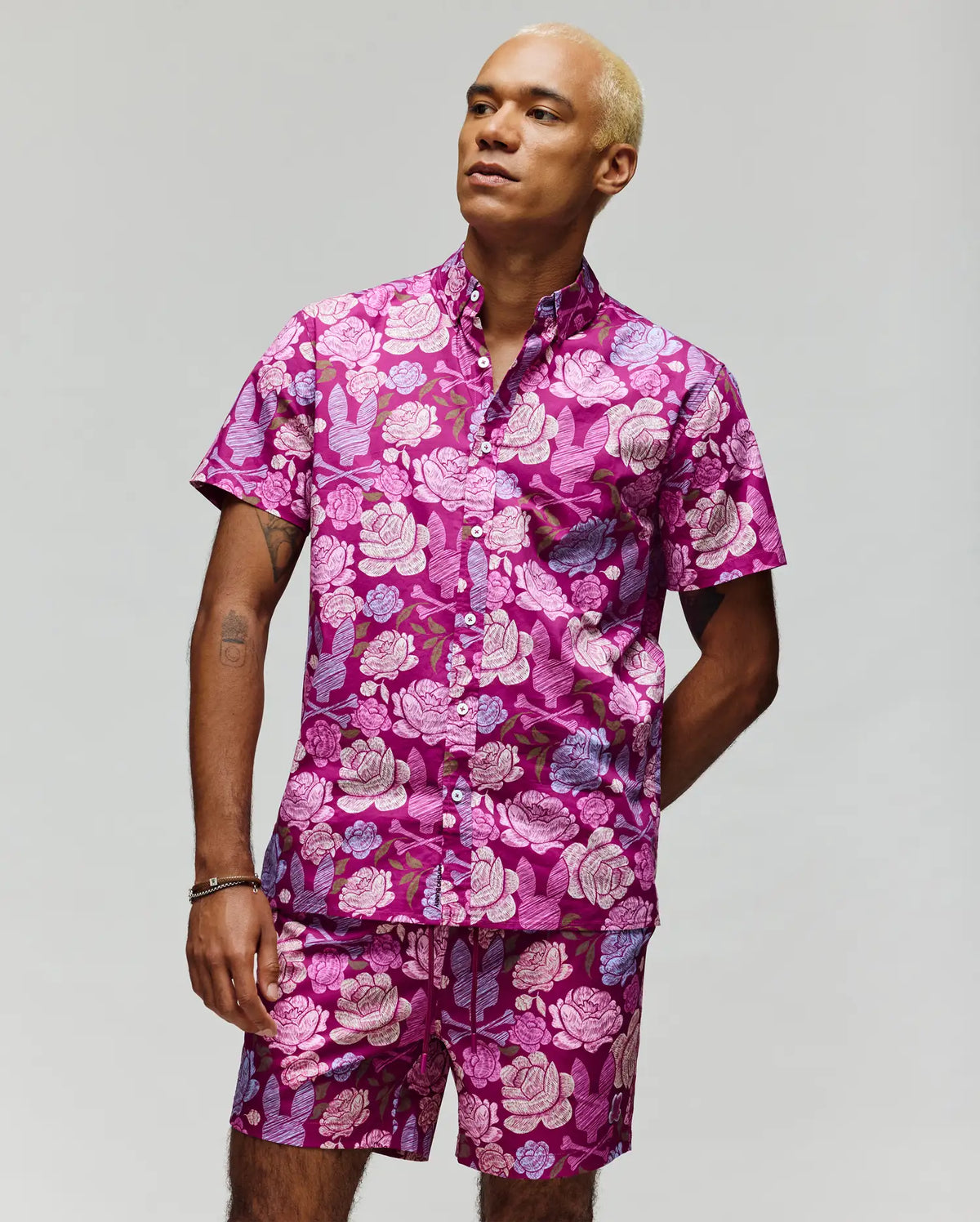 A person with short blond hair models the Psycho Bunny MENS ROWEN SHIRT - MT0300038, a bold pink, purple, and white floral print short-sleeve shirt and matching shorts, against a plain light background.