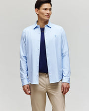 A man stands against a plain background wearing the Psycho Bunny MENS JIM JERSEY STRIPED KNIT SHIRT (MT0300041) over a navy t-shirt and beige pants, looking slightly to the side with a neutral expression.