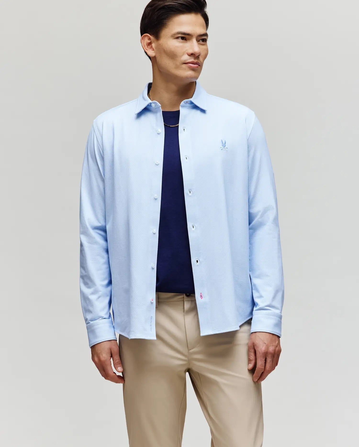 A man stands against a plain background wearing the Psycho Bunny MENS JIM JERSEY STRIPED KNIT SHIRT (MT0300041) over a navy t-shirt and beige pants, looking slightly to the side with a neutral expression.