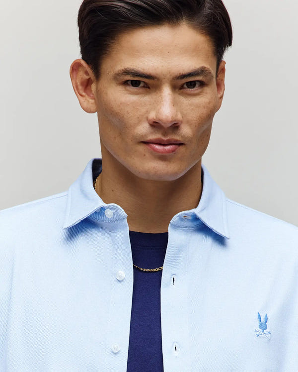 A man with short dark hair confidently models the Psycho Bunny MENS JIM JERSEY STRIPED KNIT SHIRT - MT0300041 over a navy tee and gold chain, looking at the camera against a neutral background.
