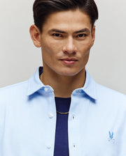 A man with short dark hair confidently models the Psycho Bunny MENS JIM JERSEY STRIPED KNIT SHIRT - MT0300041 over a navy tee and gold chain, looking at the camera against a neutral background.