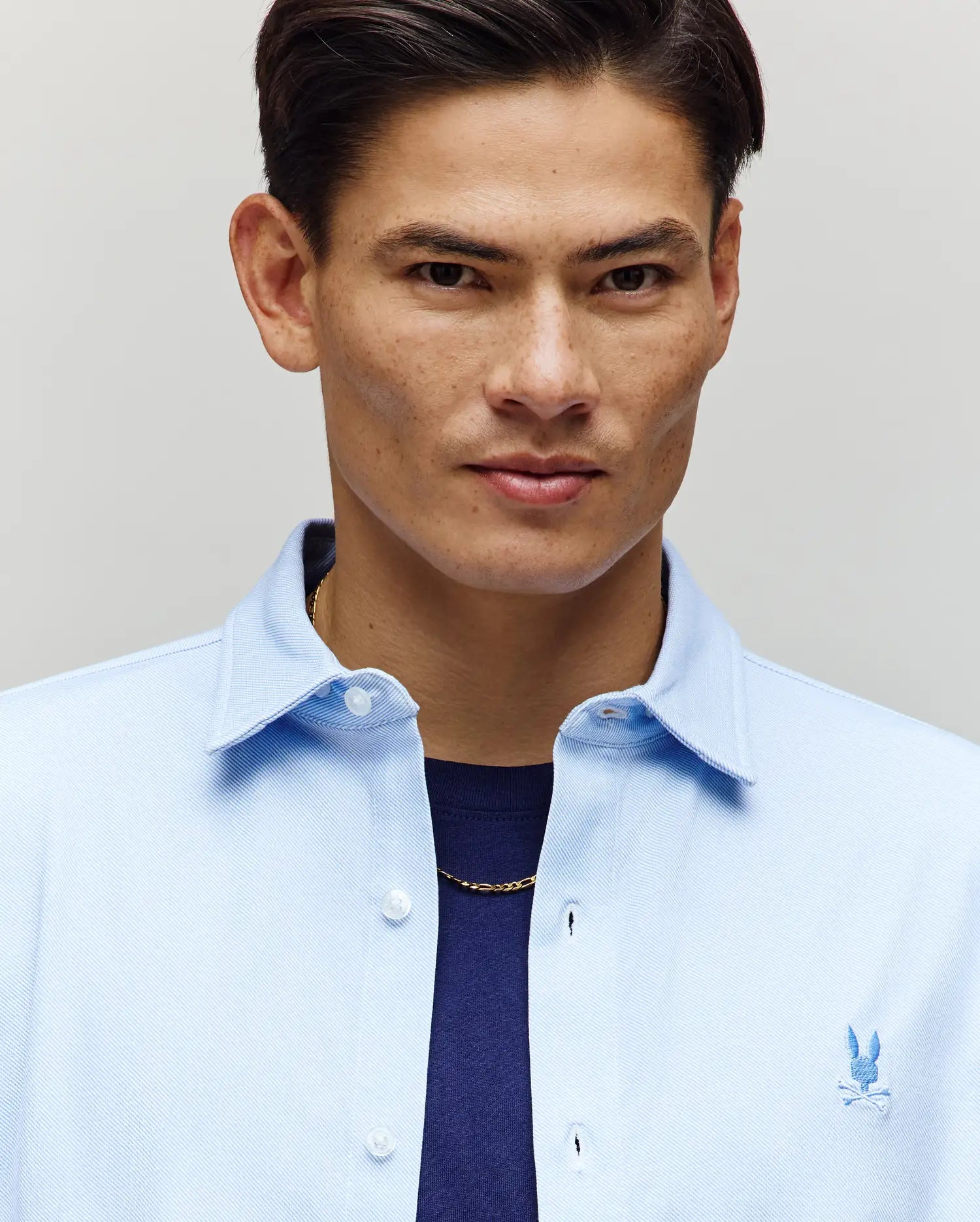A man with short dark hair confidently models the Psycho Bunny MENS JIM JERSEY STRIPED KNIT SHIRT - MT0300041 over a navy tee and gold chain, looking at the camera against a neutral background.