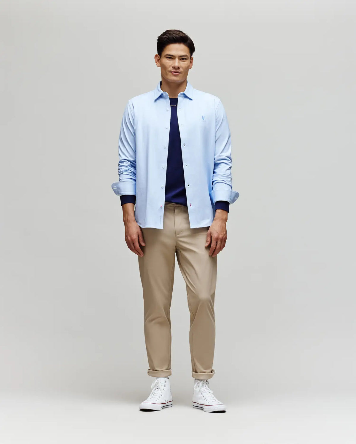 A man faces forward in a Psycho Bunny MENS JIM JERSEY STRIPED KNIT SHIRT (MT0300041), layered over a navy tee with beige pants and white sneakers, set against a plain light gray background.