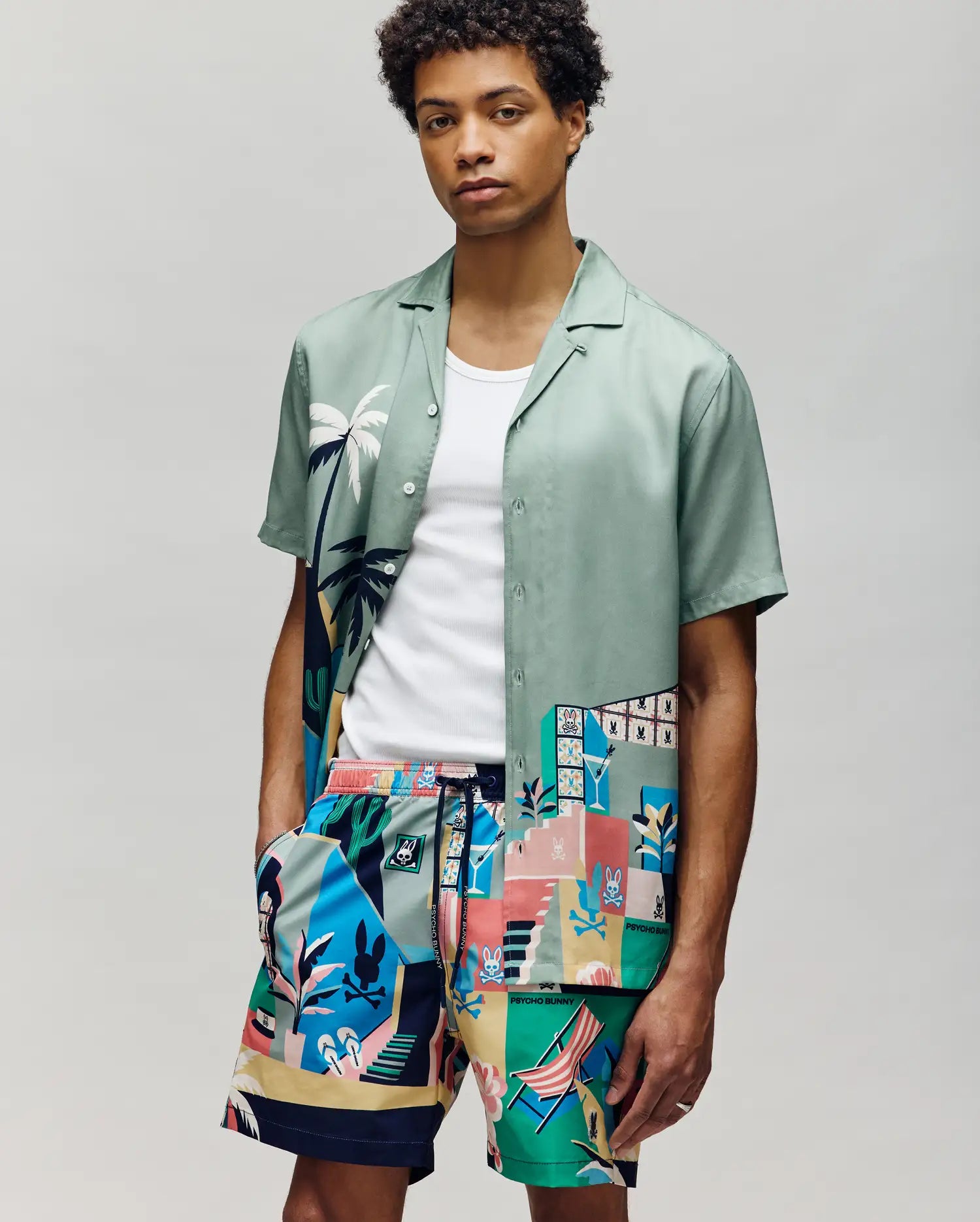 A man stands against a plain background wearing a light green Psycho Bunny MENS BLAISE TENCEL SHIRT (MT0300056) over a white tank top and tropical geometric patterned shorts, with one hand in his pocket.