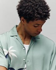A model with curly dark hair looks down, wearing the Psycho Bunny MENS BLAISE TENCEL SHIRT - MT0300056 in light green with a white palm tree print over a white undershirt, set against a plain light background.