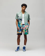 A man in a breathable light green Psycho Bunny MENS BLAISE TENCEL SHIRT (MT0300056), white tank top, tropical abstract shorts, and blue-white high-top sneakers stands against a plain gray background.