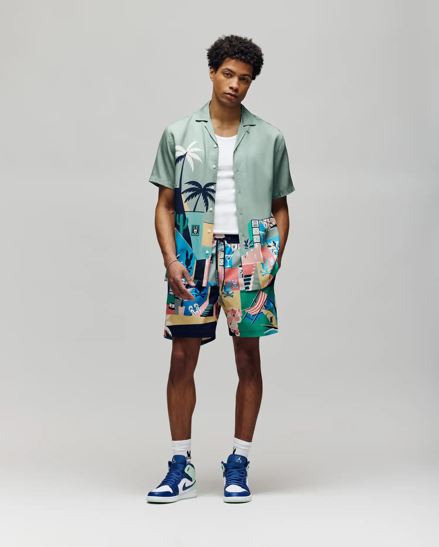 A man in a breathable light green Psycho Bunny MENS BLAISE TENCEL SHIRT (MT0300056), white tank top, tropical abstract shorts, and blue-white high-top sneakers stands against a plain gray background.