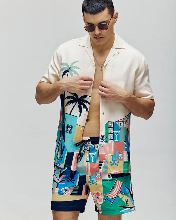 A man in sunglasses and a colorful Psycho Bunny MENS BLAISE TENCEL SHIRT (MT0300056) with matching tropical swim trunks stands against a light background, partially unbuttoning his 100% Tencel shirt.