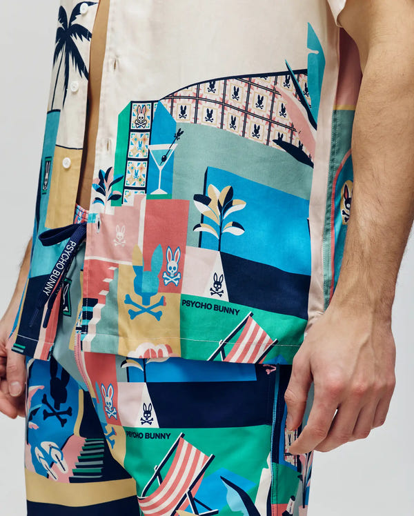 A person wears the MENS BLAISE TENCEL SHIRT - MT0300056 by Psycho Bunny, a colorful short-sleeve button-up made from 100% Tencel, featuring abstract beach and bunny motifs in blue, pink, green, and beige.