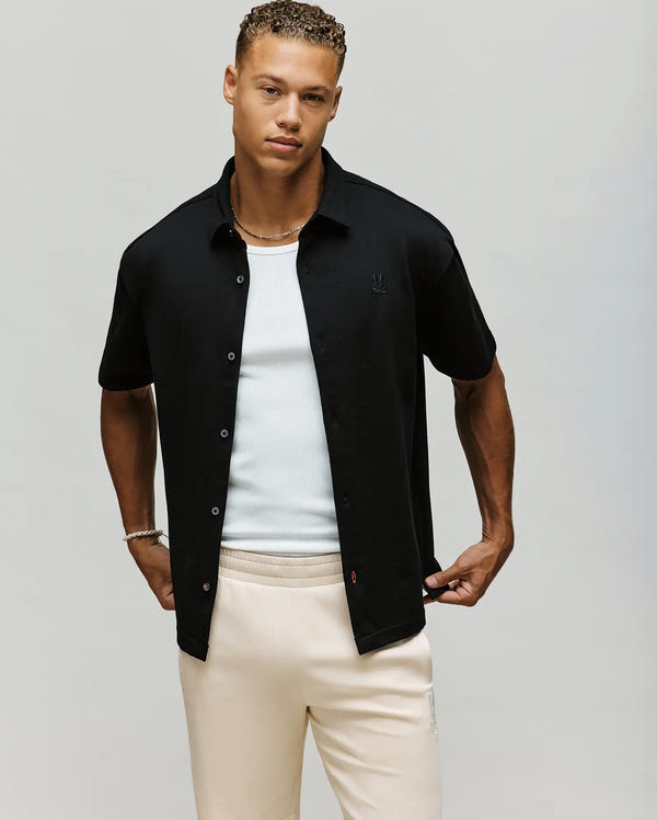 A young man wears the Psycho Bunny MENS KIAN OVERSIZED FIT SHIRT - MT0300063, a relaxed black short-sleeve button-up with mother-of-pearl buttons, styled over a white ribbed tank and light beige pants, posing against a plain background.