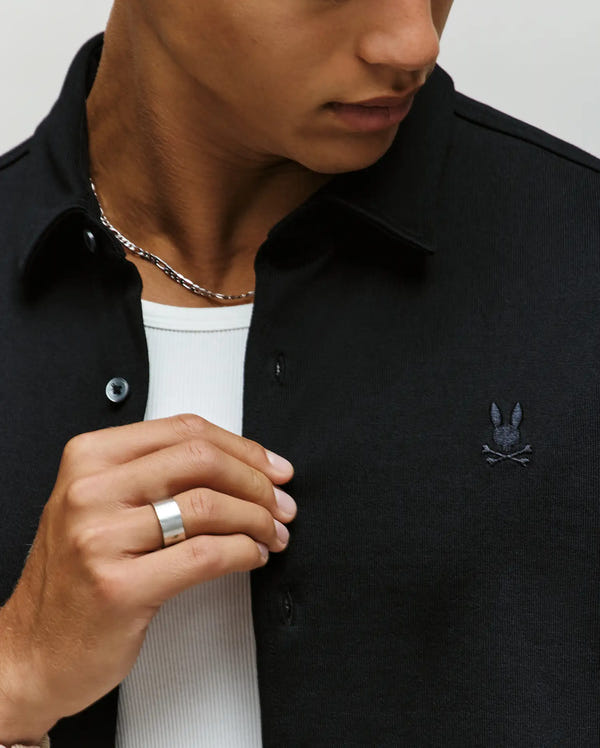 A person wearing the Psycho Bunny MENS KIAN OVERSIZED FIT SHIRT (MT0300063)—a black collared shirt with a small embroidered logo and mother-of-pearl buttons—layered over a white ribbed shirt, accessorized with a silver chain necklace and ring.