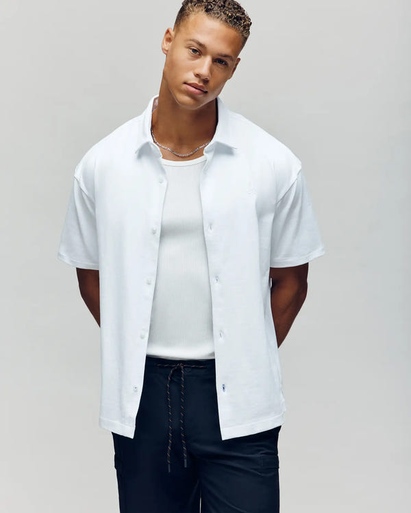 A young man with short curly hair wears the Psycho Bunny MENS KIAN OVERSIZED FIT SHIRT (MT0300063) in white over a ribbed tank top and black pants, standing with hands behind his back against a plain light background.