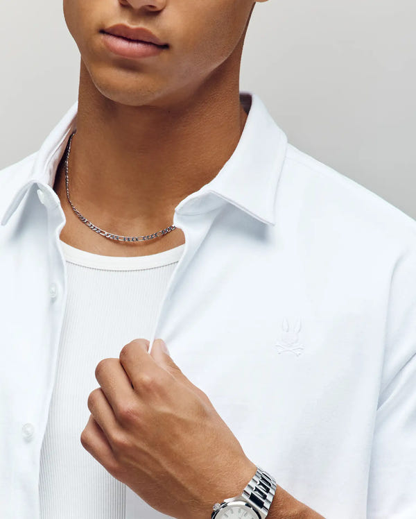 A person wears the Psycho Bunny MENS KIAN OVERSIZED FIT SHIRT (MT0300063) in white, unbuttoned over a white ribbed tank top, accessorized with a silver chain necklace and watch. The background is plain and neutral.