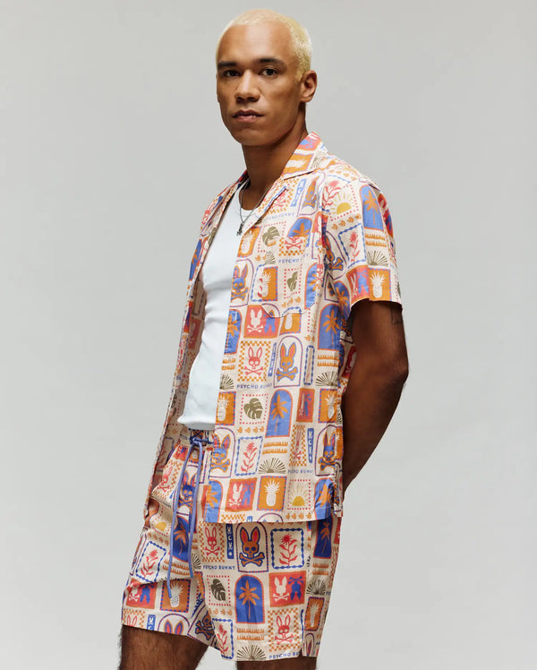 A person with short blond hair wears the Psycho Bunny MENS SUNDEW SHIRT (MT0300064), a colorful, patterned cotton shirt over a white t-shirt, paired with matching shorts, standing against a plain light background.