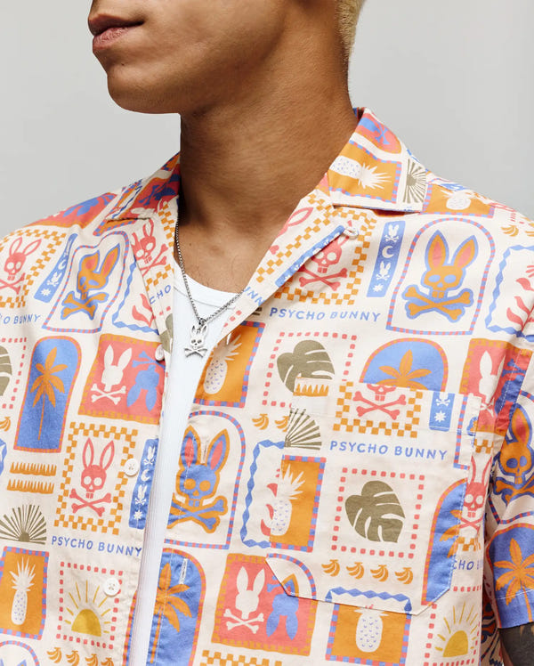 A person wearing the Psycho Bunny MENS SUNDEW SHIRT - MT0300064, a colorful short-sleeve cotton shirt with playful orange, blue, and pink patterns, layered over a white shirt and accessorized with a silver necklace.