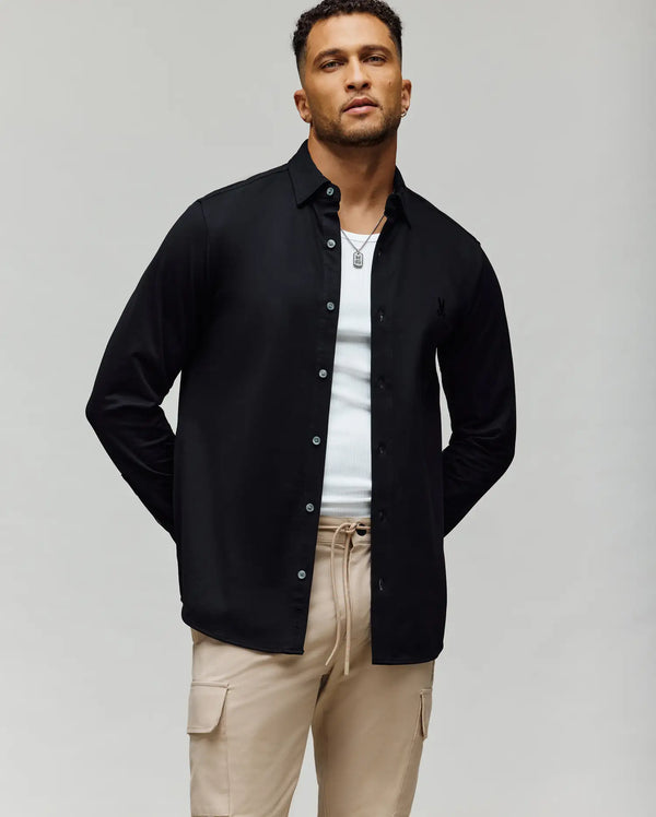 A man stands against a plain background wearing the Psycho Bunny MENS JIM JERSEY KNIT SHIRT - MT0300067 over a white tank top, with beige cargo pants and a necklace. His hands are behind his back and he is looking slightly upward.