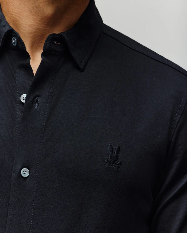 A close-up of a person wearing the Psycho Bunny MENS JIM JERSEY KNIT SHIRT (MT0300067), a dark athletic-fit, long-sleeve button-up with light-colored buttons, pointed collar, and subtle rabbit and crossbones chest embroidery.