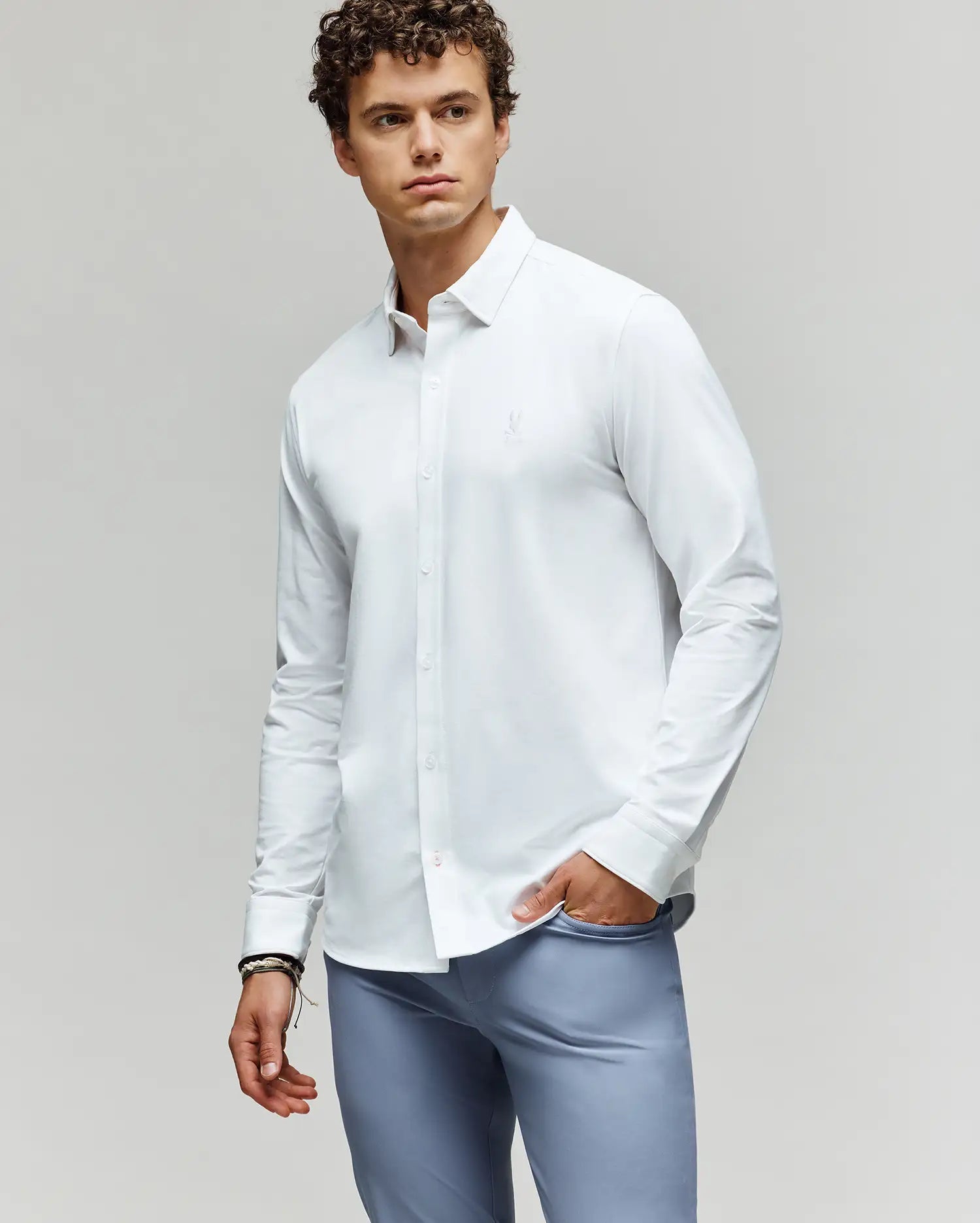 A young man with curly hair wears the Psycho Bunny MENS JIM JERSEY KNIT SHIRT (MT0300067) and light blue pants, standing against a plain background with one hand in his pocket, looking to the side with a neutral expression.
