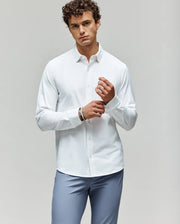 A man with curly brown hair wears the Psycho Bunny MENS JIM JERSEY KNIT SHIRT (MT0300067) in white and light blue pants, standing against a plain light gray background while adjusting his left cuff and looking at the camera.