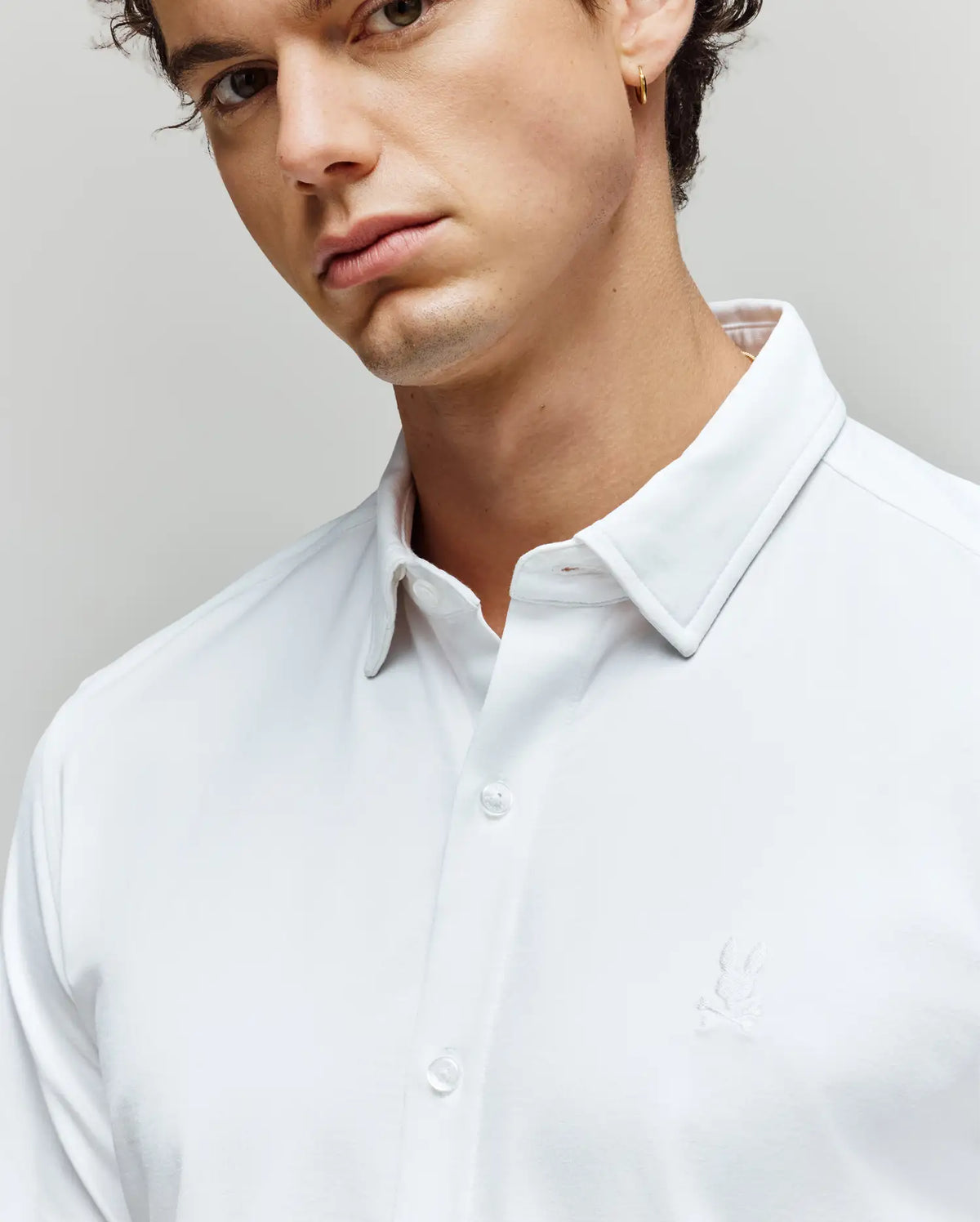 A young man wears the Psycho Bunny MENS JIM JERSEY KNIT SHIRT (MT0300067), an athletic fit white collared button-up with subtle embroidery, looking slightly to the side against a plain background.