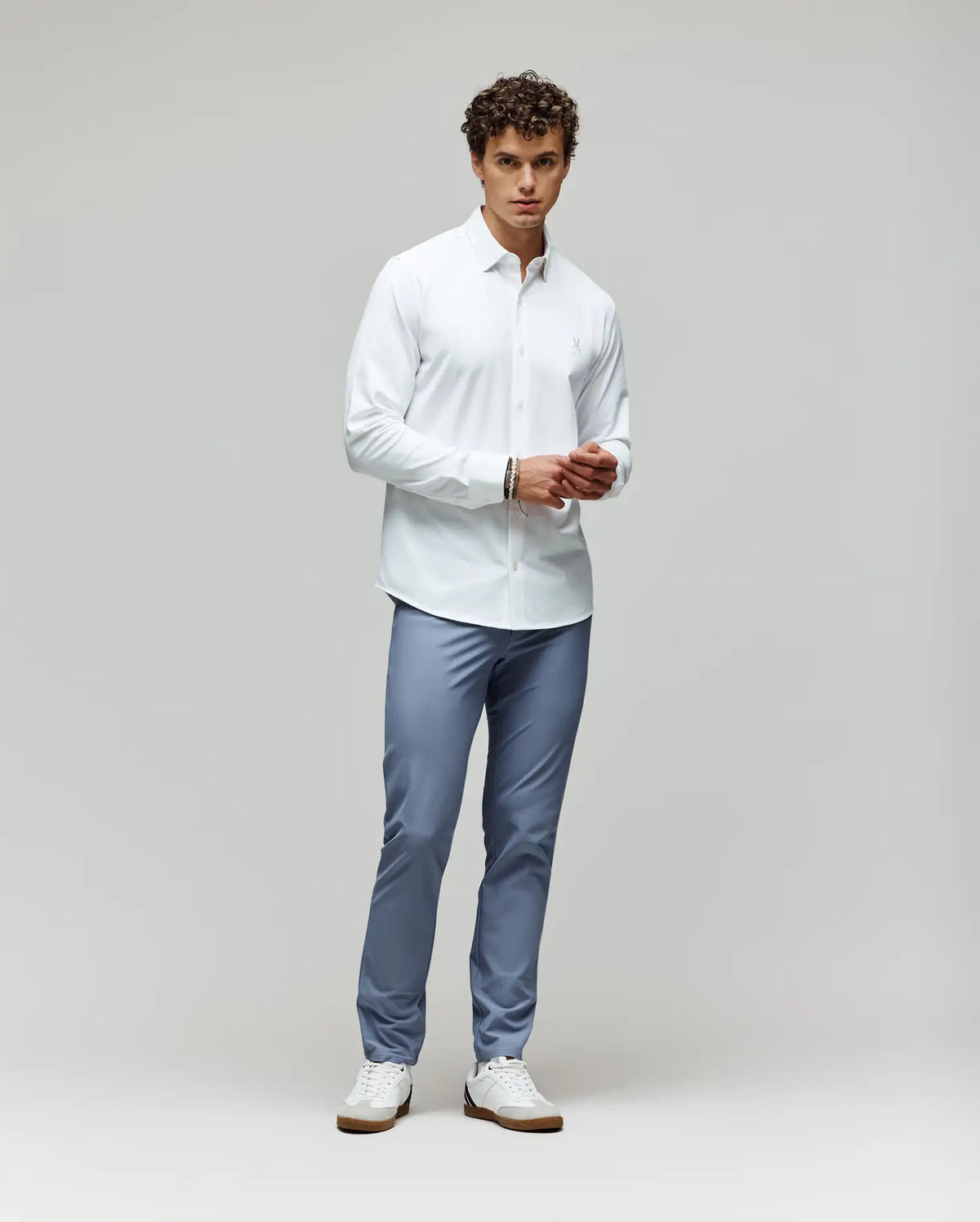 A man with curly hair stands against a plain background, wearing the Psycho Bunny MENS JIM JERSEY KNIT SHIRT (MT0300067) and light blue trousers with white sneakers, facing forward with his hands together at his waist.