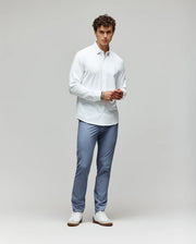 A man with curly hair stands against a plain background, wearing the Psycho Bunny MENS JIM JERSEY KNIT SHIRT (MT0300067) and light blue trousers with white sneakers, facing forward with his hands together at his waist.