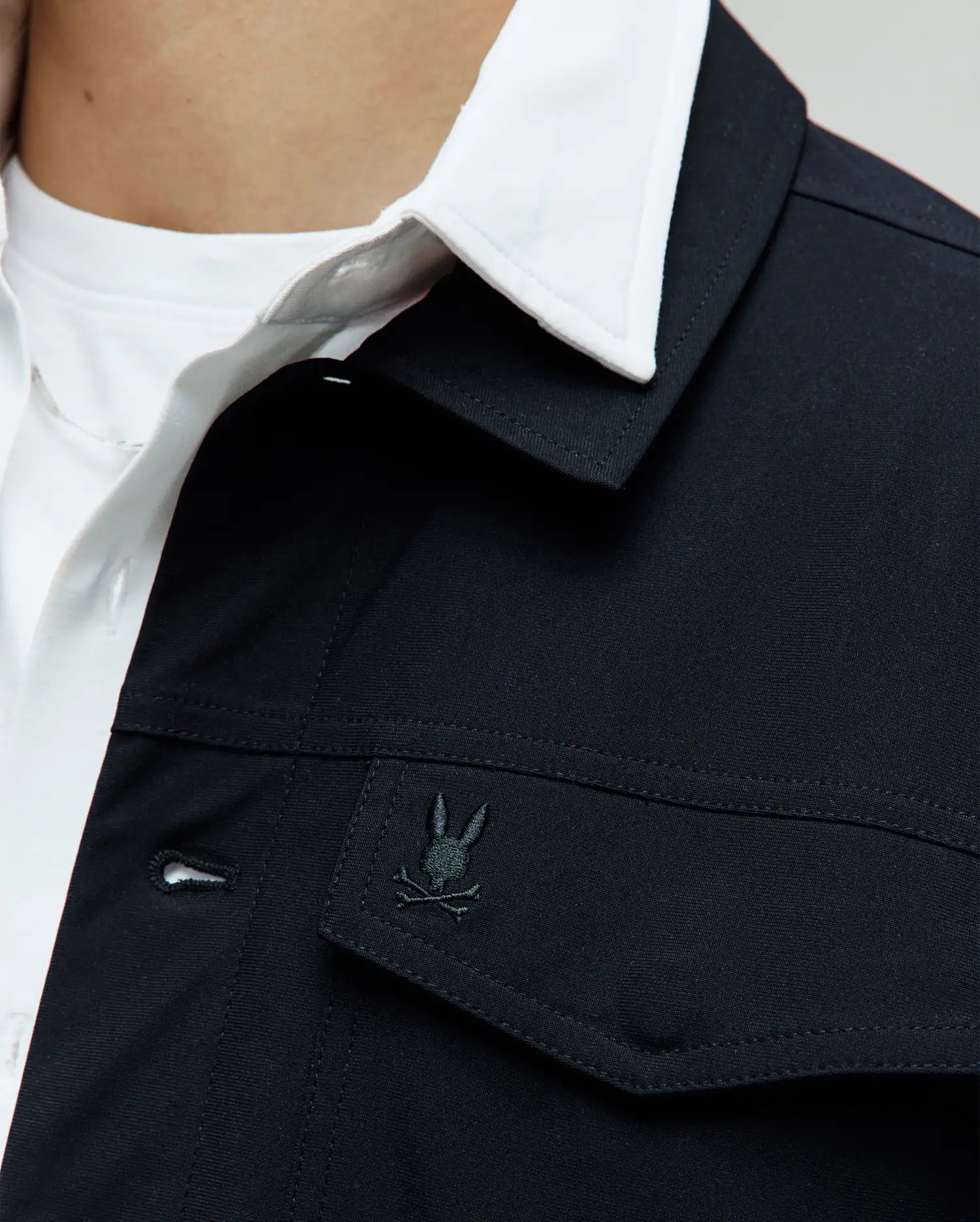 Black Mens Maverick Overshirt, a classic fit with smooth fabric, featuring a front button placket and a small embroidered Psycho Bunny skull logo on the left chest pocket flap, layered over a white collared shirt.