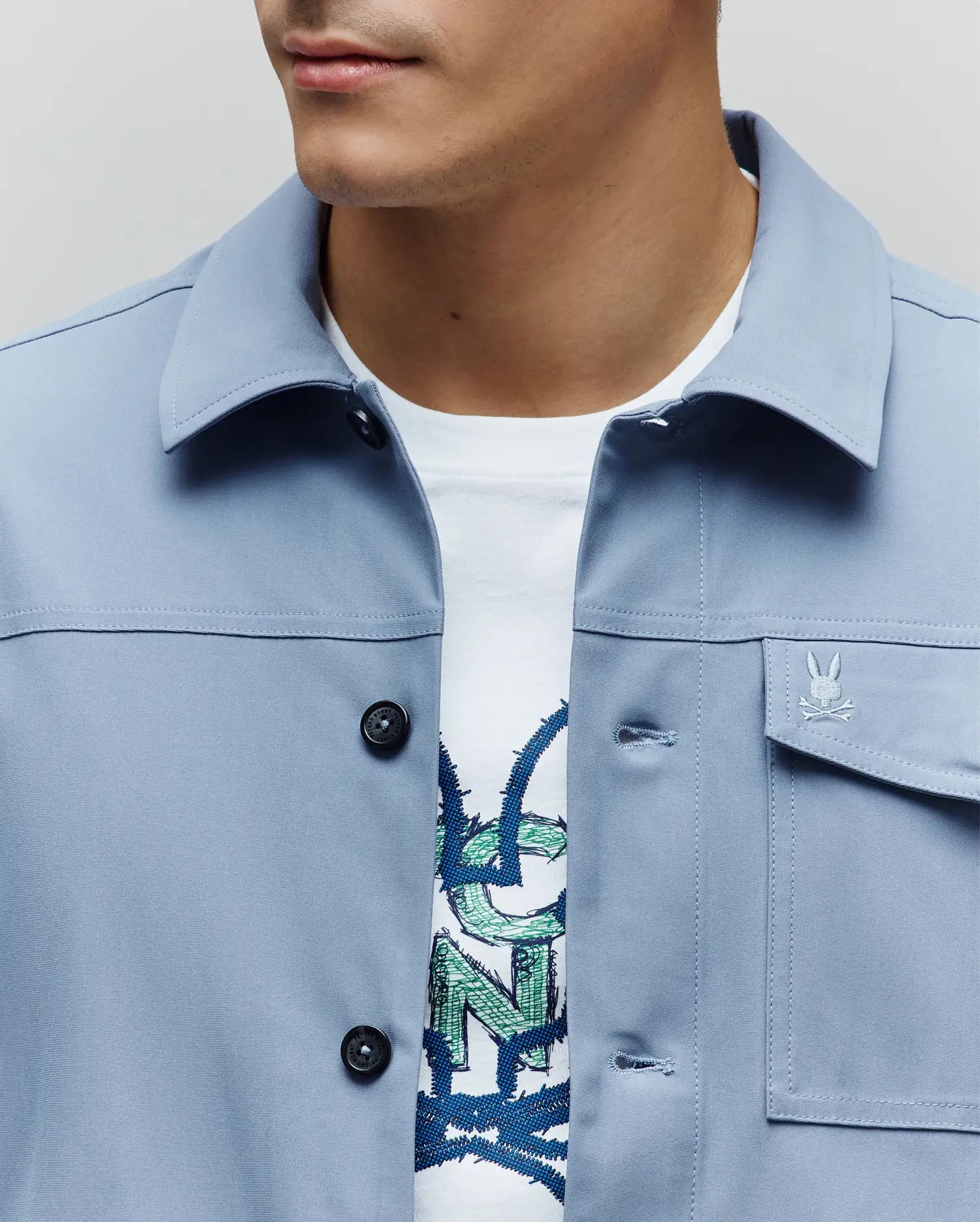 Mens Maverick Overshirt in light blue, featuring a classic fit, smooth woven fabric, black button-front closure, and a small embroidered Psycho Bunny skull logo on the right chest pocket.