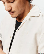 Mens Sunny Linen Shirt, a relaxed fit off-white linen camp collar shirt featuring a small embroidered Psycho Bunny skull logo on the left chest and black and teal chevron sleeve pattern. (2)