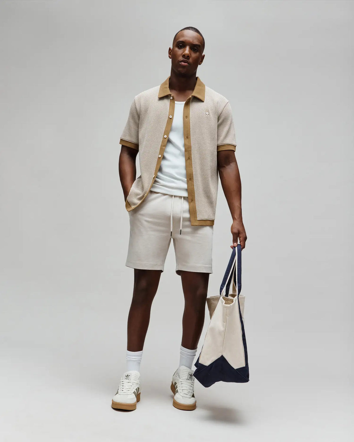 Mens Maxton Waffle Shirt, a beige waffle knit polo with tan accents on the collar and cuffs, featuring a small embroidered Psycho Bunny skull logo on the chest, paired with light cream drawstring shorts and a light blue t-shirt.