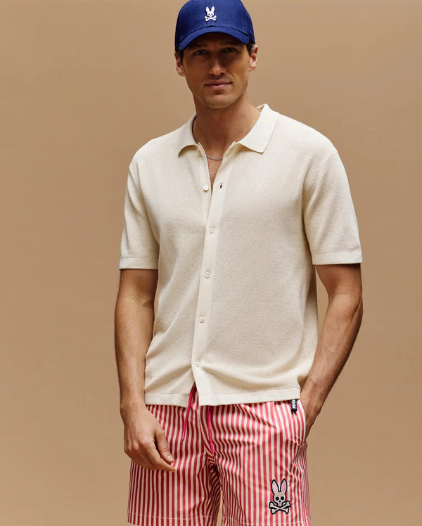 A man wears a blue cap, Psycho Bunny MENS MORGAN TEXTURED KNIT SHIRT (MT0400002) in cream, and red-white striped shorts with a cartoon bunny logo, standing against a plain beige background.