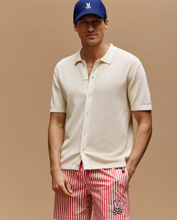 A man wears a blue cap, Psycho Bunny MENS MORGAN TEXTURED KNIT SHIRT (MT0400002) in cream, and red-white striped shorts with a cartoon bunny logo, standing against a plain beige background.