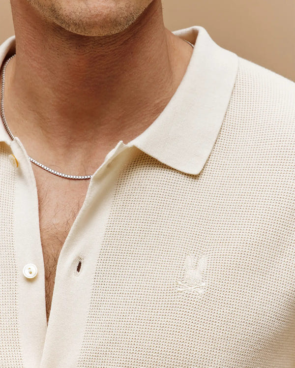 Close-up of a person wearing the Psycho Bunny MENS MORGAN TEXTURED KNIT SHIRT (MT0400002), cream-colored with buttons undone, subtle chest logo embroidery, and paired with a thin silver chain necklace.