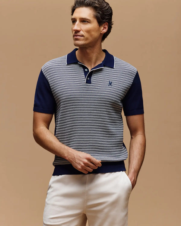 A man wears the Psycho Bunny MENS KENNY STRIPED KNIT POLO - MT0400003, a short-sleeve navy and white striped classic style with breathable fabric, paired with light-colored pants, standing against a solid beige background.