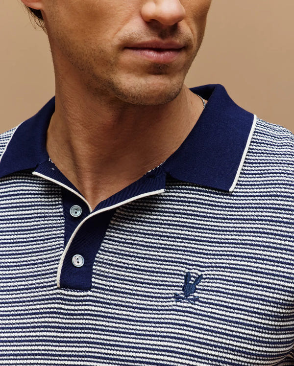 A man wears the Psycho Bunny MENS KENNY STRIPED KNIT POLO (MT0400003), featuring navy and white stripes, a dark blue collar with white trim, breathable fabric, and a small chest logo. His face is partially visible.