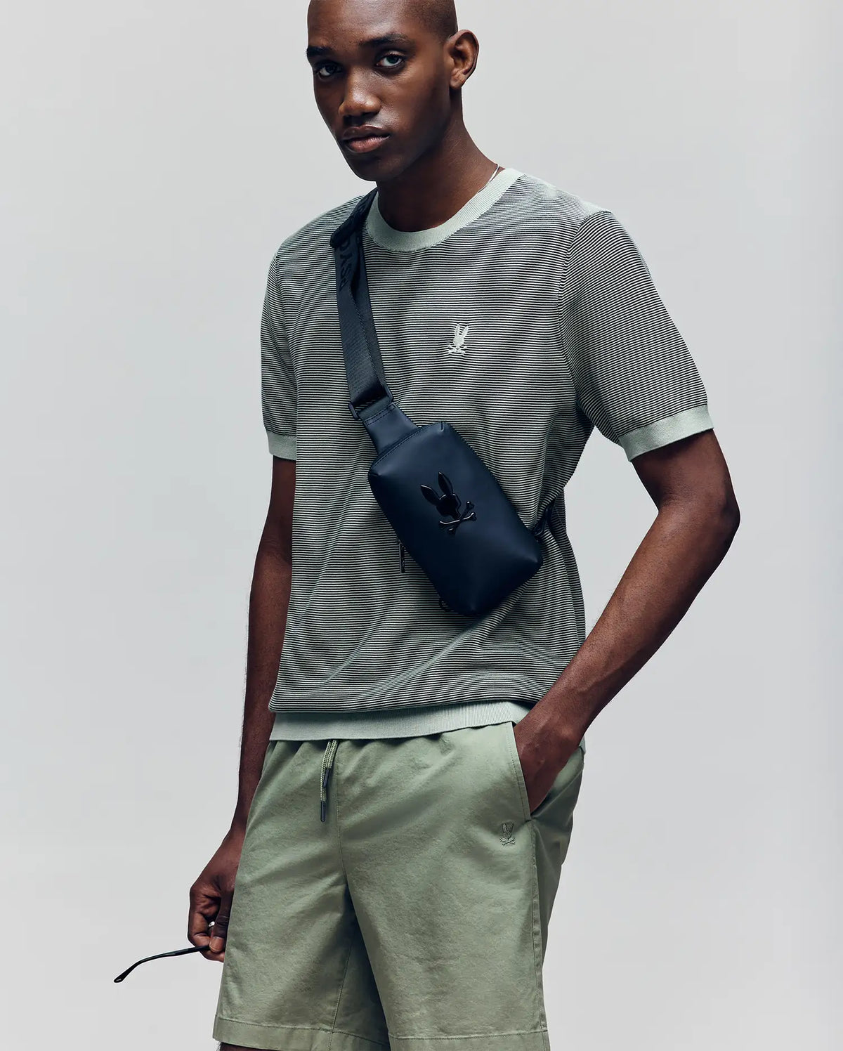 A man wears a sage green Psycho Bunny MENS MARIO STRIPED SWEATER T-SHIRT (MT0400009) with matching shorts and a black crossbody bag featuring an embroidered bunny logo, standing against a plain light gray background.